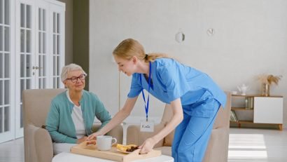 Positive young nurse carries tray with breakfast to senior woman patient sitting in light living room