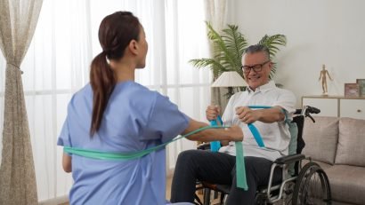 selective focus of asian senior disabled man strengthening his bicep by doing physiotherapy resistance training using elastic band. he sits on wheelchair with his care attendant at home